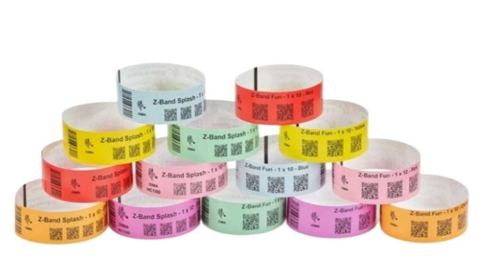 Stacked rolls of Zebra Z-Band Fun wristbands in bright colours, including red, orange, yellow, green, blue, pink, and purple, featuring QR codes and space for printable information, designed for event and patient identification.